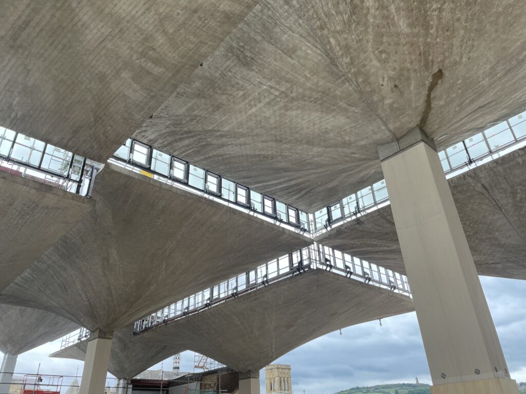 Interior view of a modern concrete building under construction, showcasing large, sculptural concrete roof slabs supported by columns. The slabs have a geometric, folded plate design with clerestory windows allowing natural light to enter. Scaffolding and construction materials are visible, and a historic tower and green hills can be seen in the background.